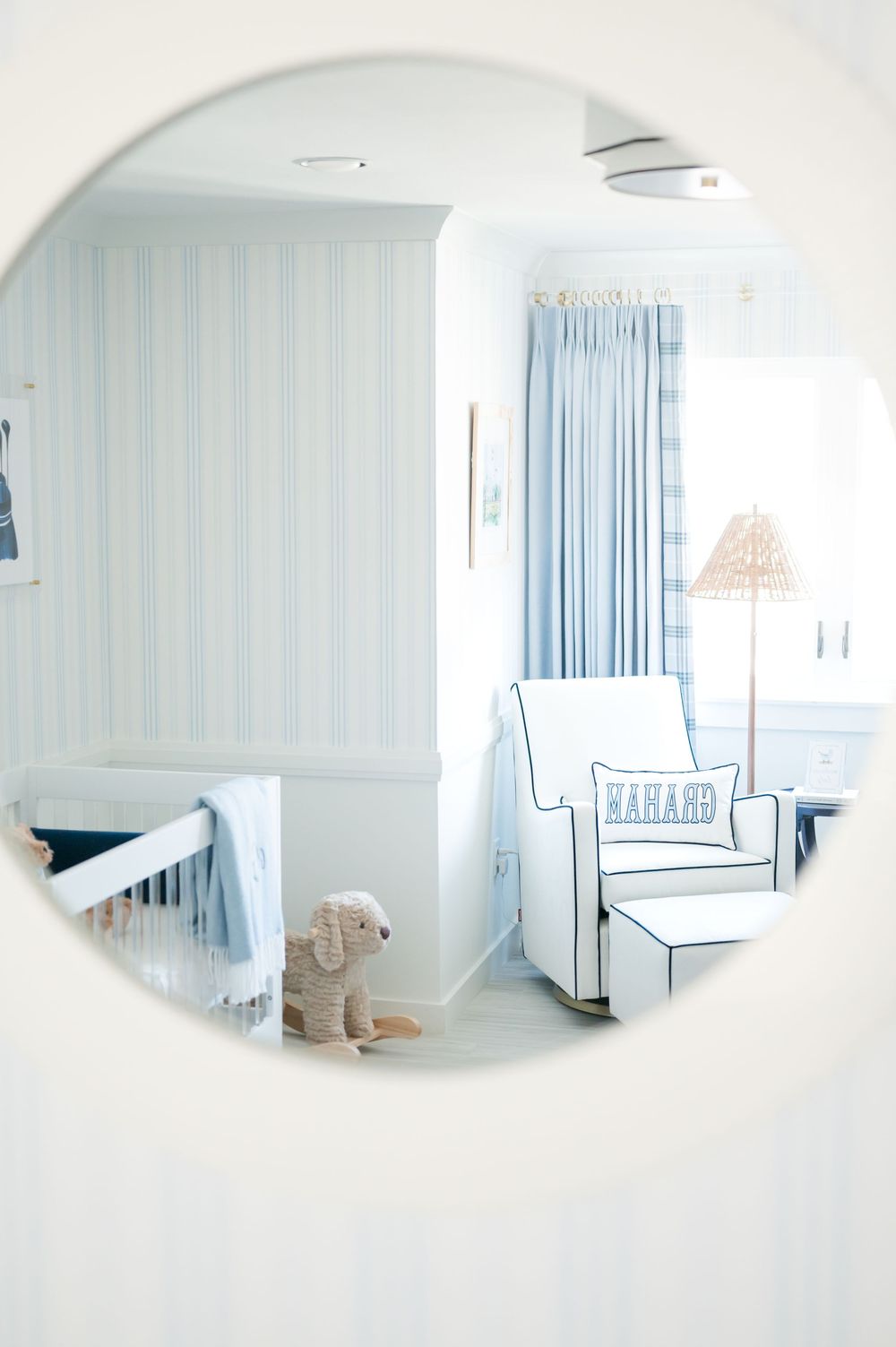 Round mirror reflection showing a serene nursery with white rocking chair, blue curtains, and soft pastel decor.