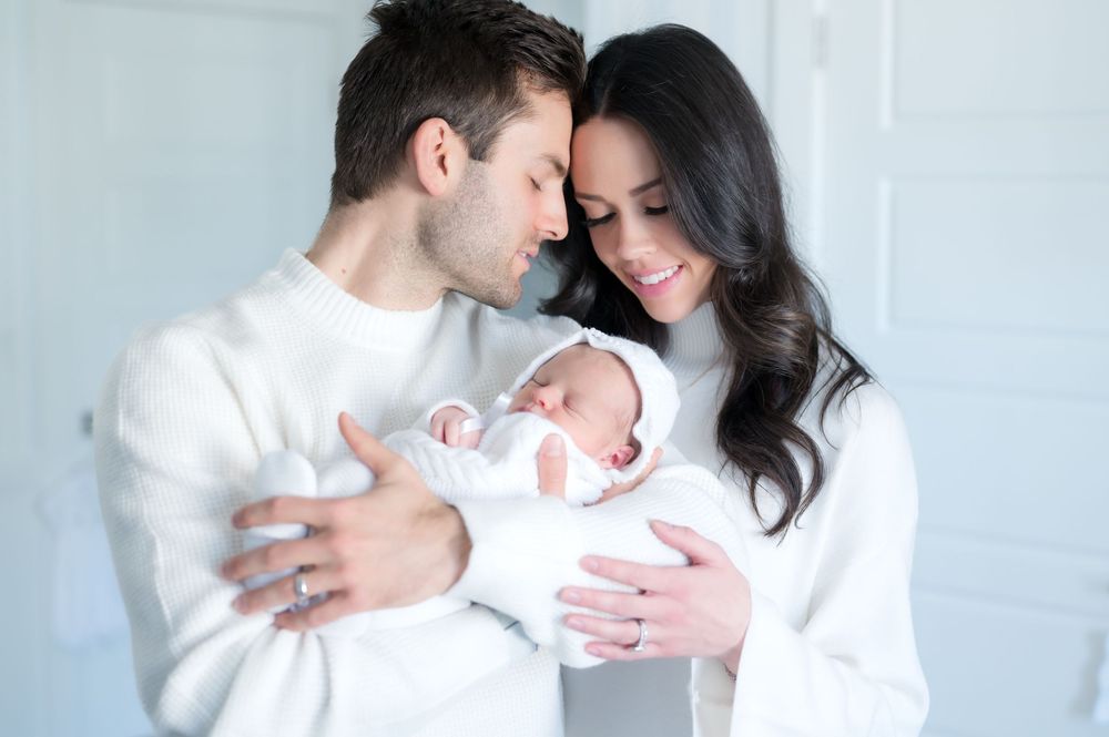Two people in white outfits tenderly hold their newborn baby in a bright minimalist setting.