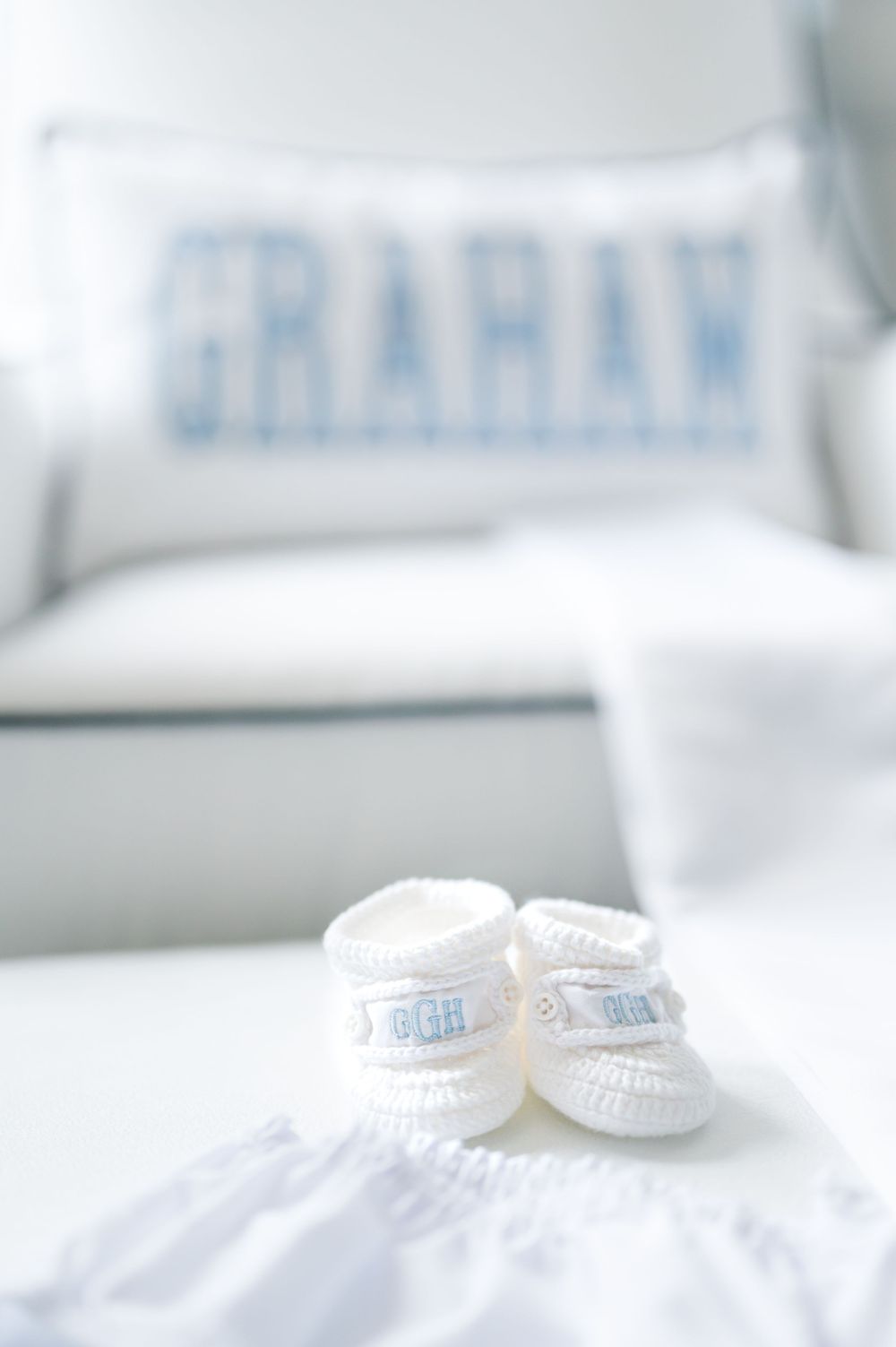 Tiny white knitted baby booties rest on soft white fabric with a blurred pillow in the background.