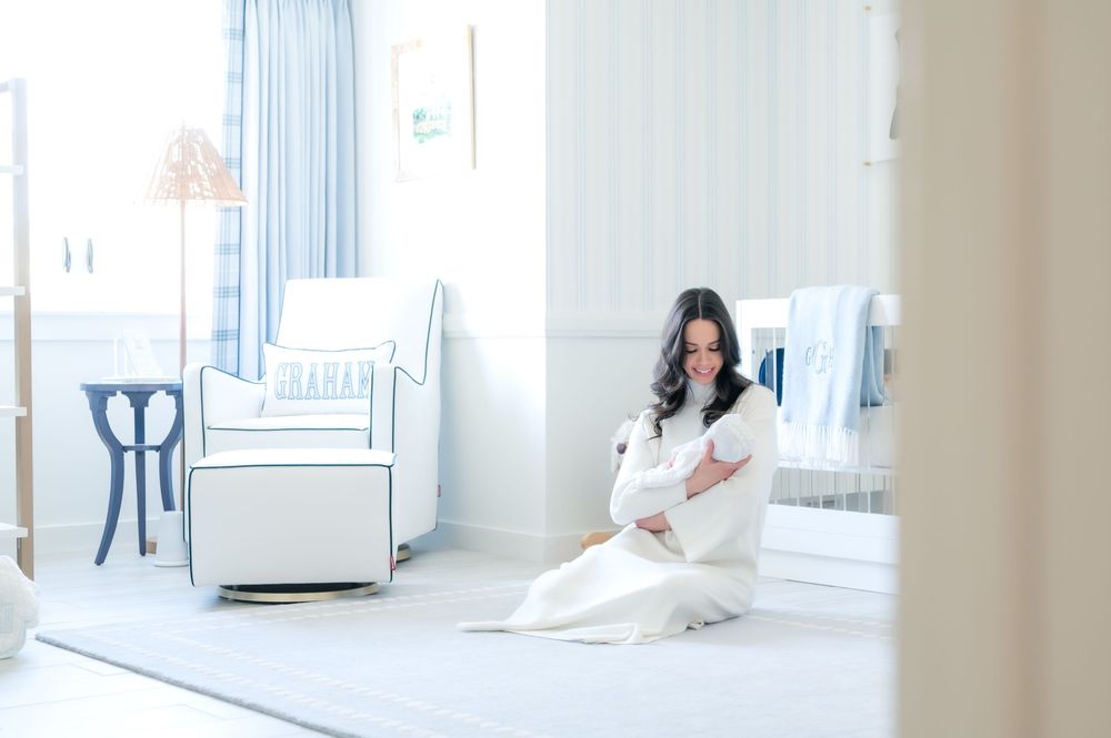 Woman in white dress sits peacefully on floor of serene nursery with rocking chair and blue curtains.