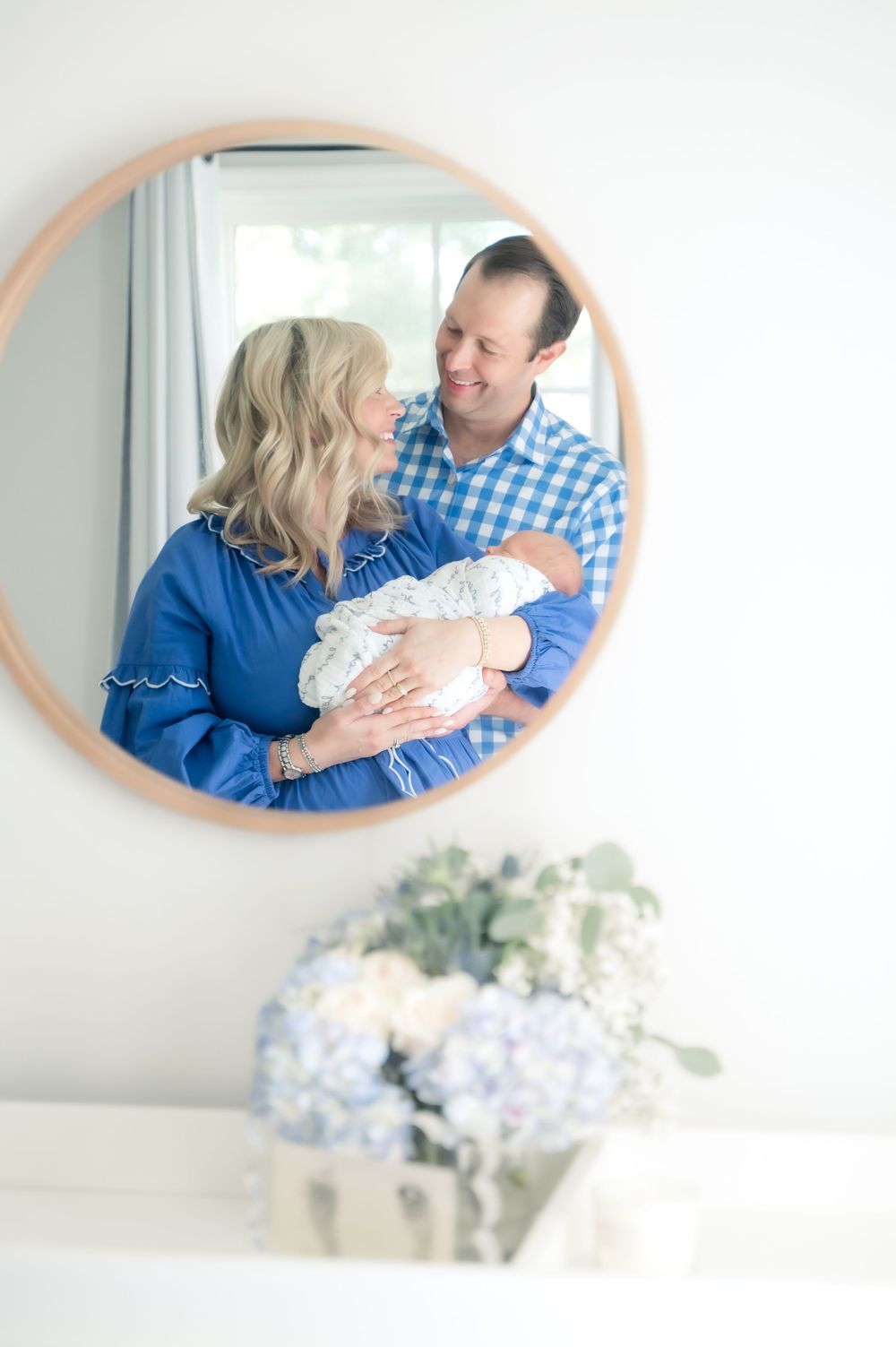 A couple in coordinating blue outfits holds their newborn baby while being reflected in a round mirror above floral decor.