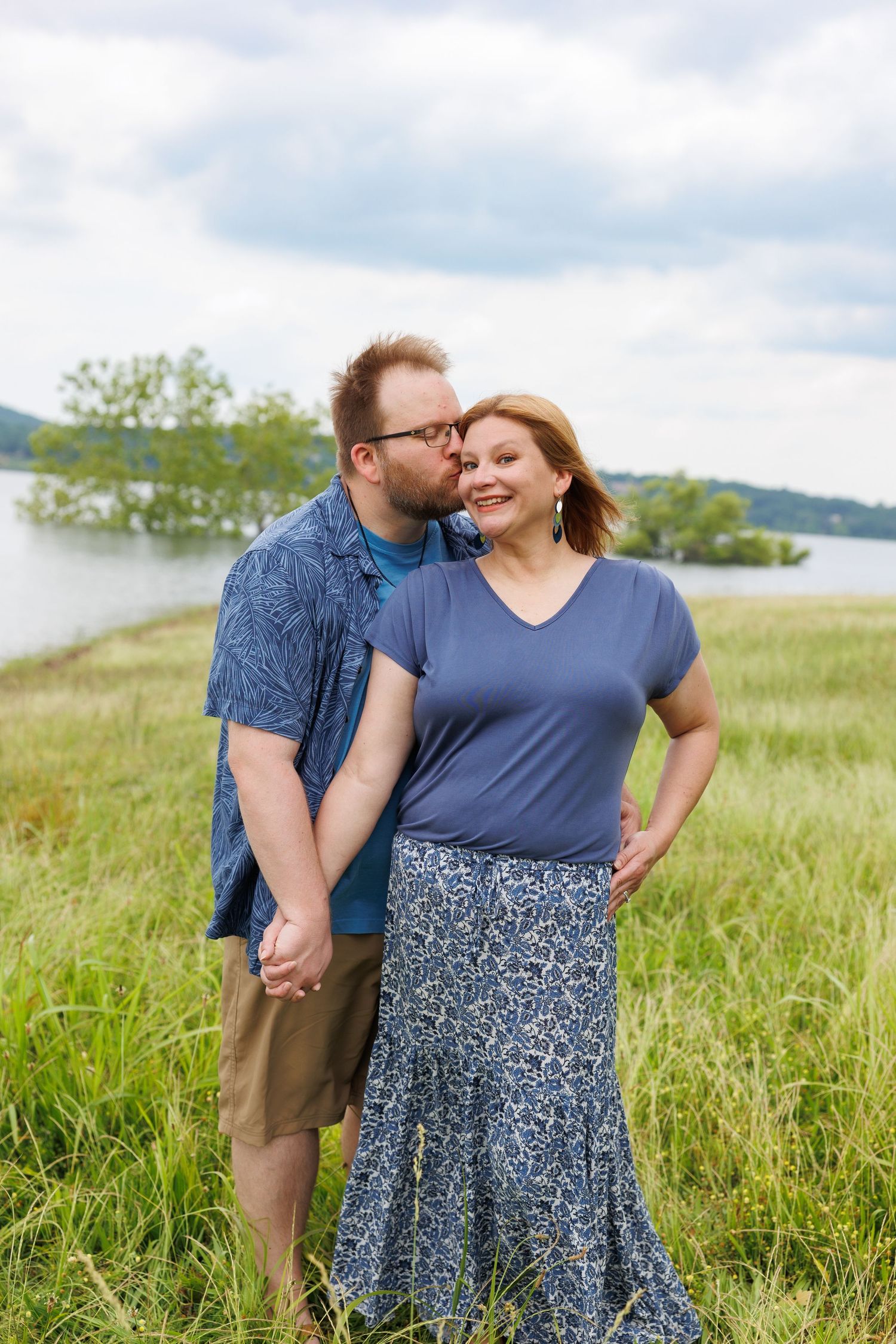 Affectionate couple shares tender moment in field of tall grass beside waterfront, with mountains in background.
