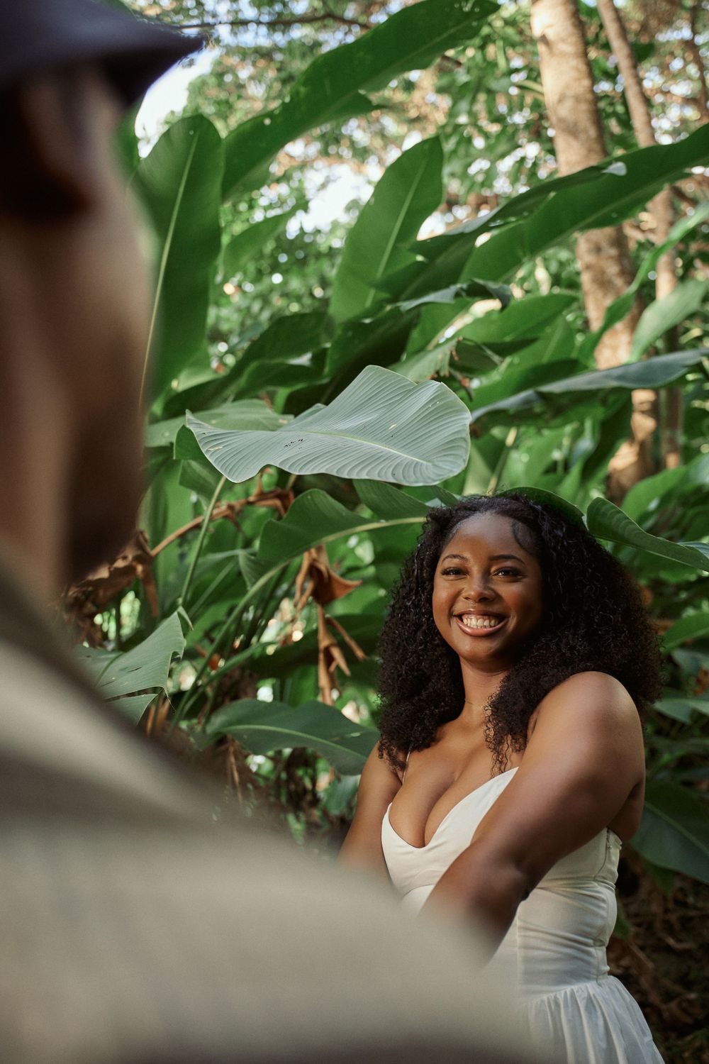 Person in white dress poses gracefully among lush green foliage in a garden setting, creating a Jamaican Photographer