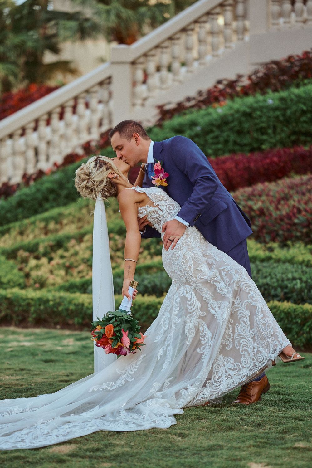 A romantic wedding couple shares a kiss on a manicured lawn with ornate staircase and colorful Jamaica Wedding Photographer