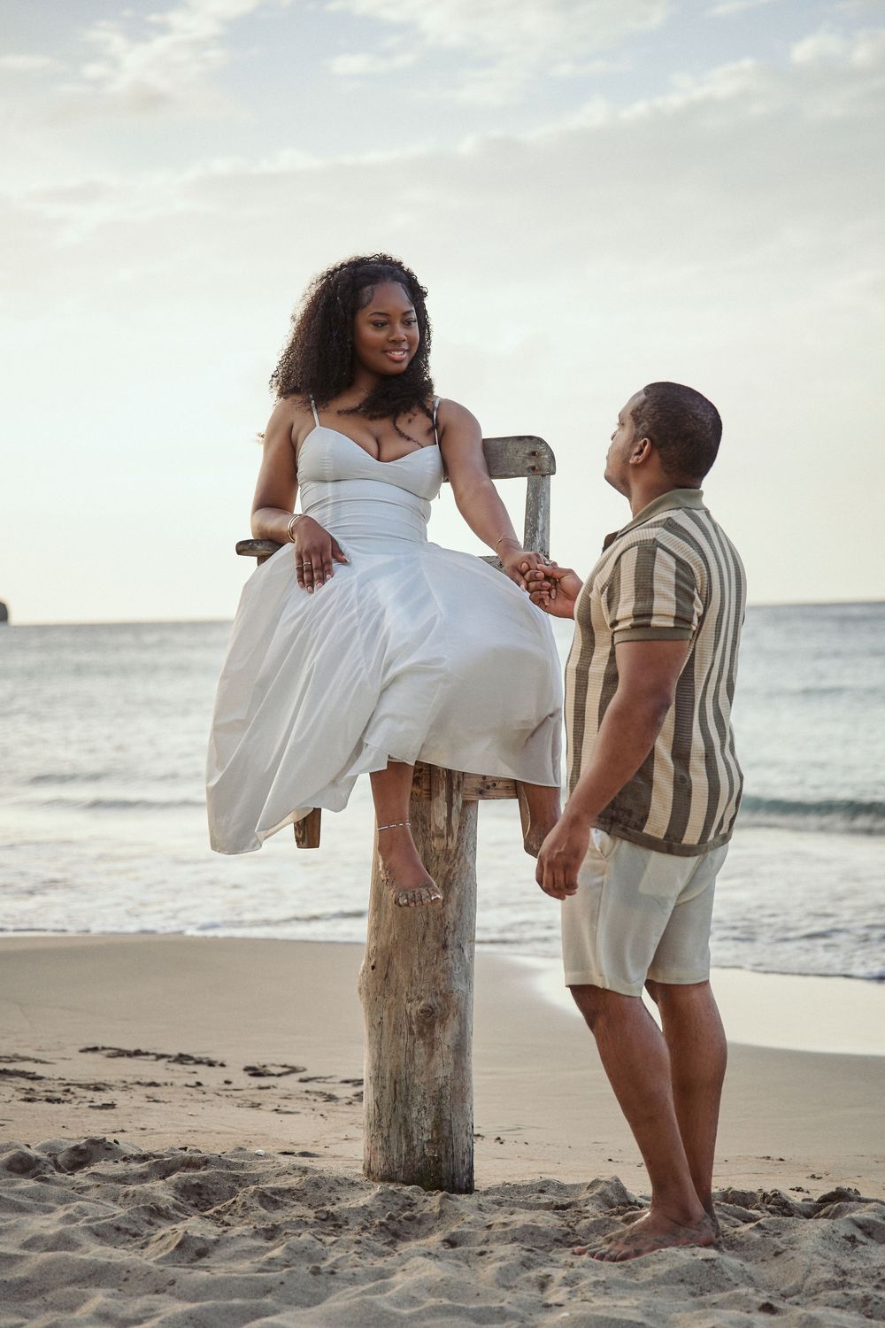 A joyful couple shares a romantic moment on a beach at sunset as she sits playfully on a wooden post. Jamaican Photographer