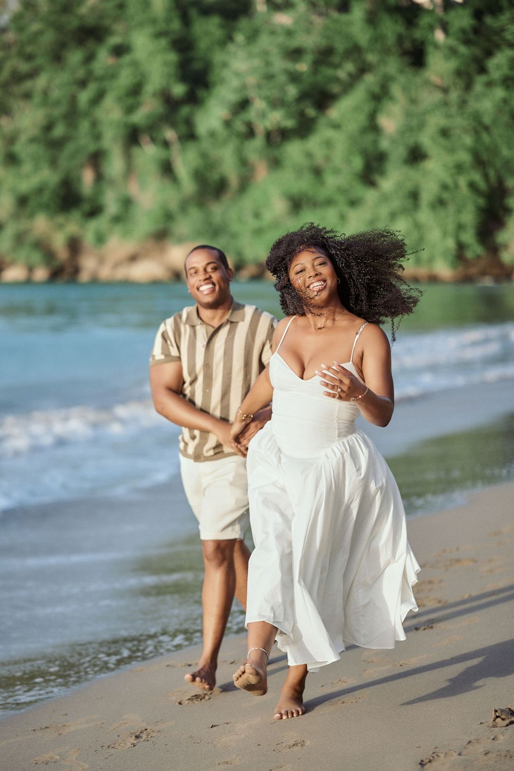 A couple runs joyfully along a tropical beach with waves and lush greenery in the background. Jamaican Photographer