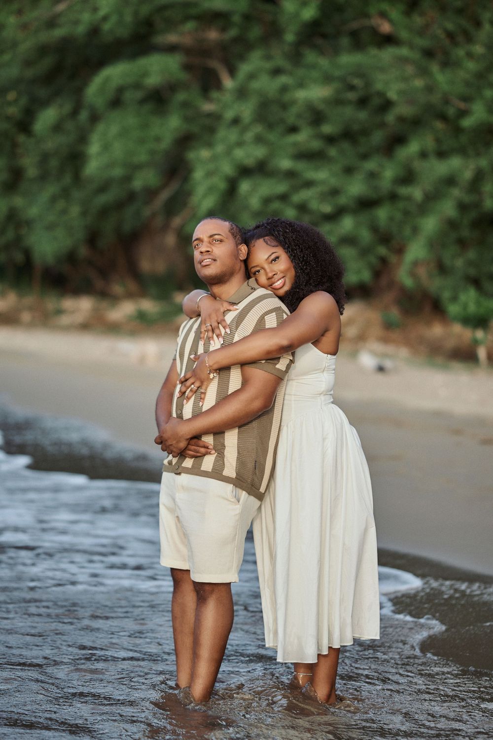 Two figures embrace joyfully while standing in shallow ocean waters at sunset on a sandy beach. Jamaican Photographer