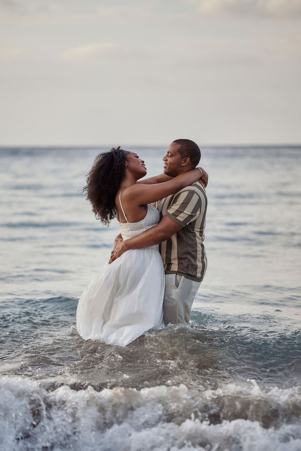A romantic couple embraces while standing in ocean waves at sunset, with her white dress Jamaican Photographer