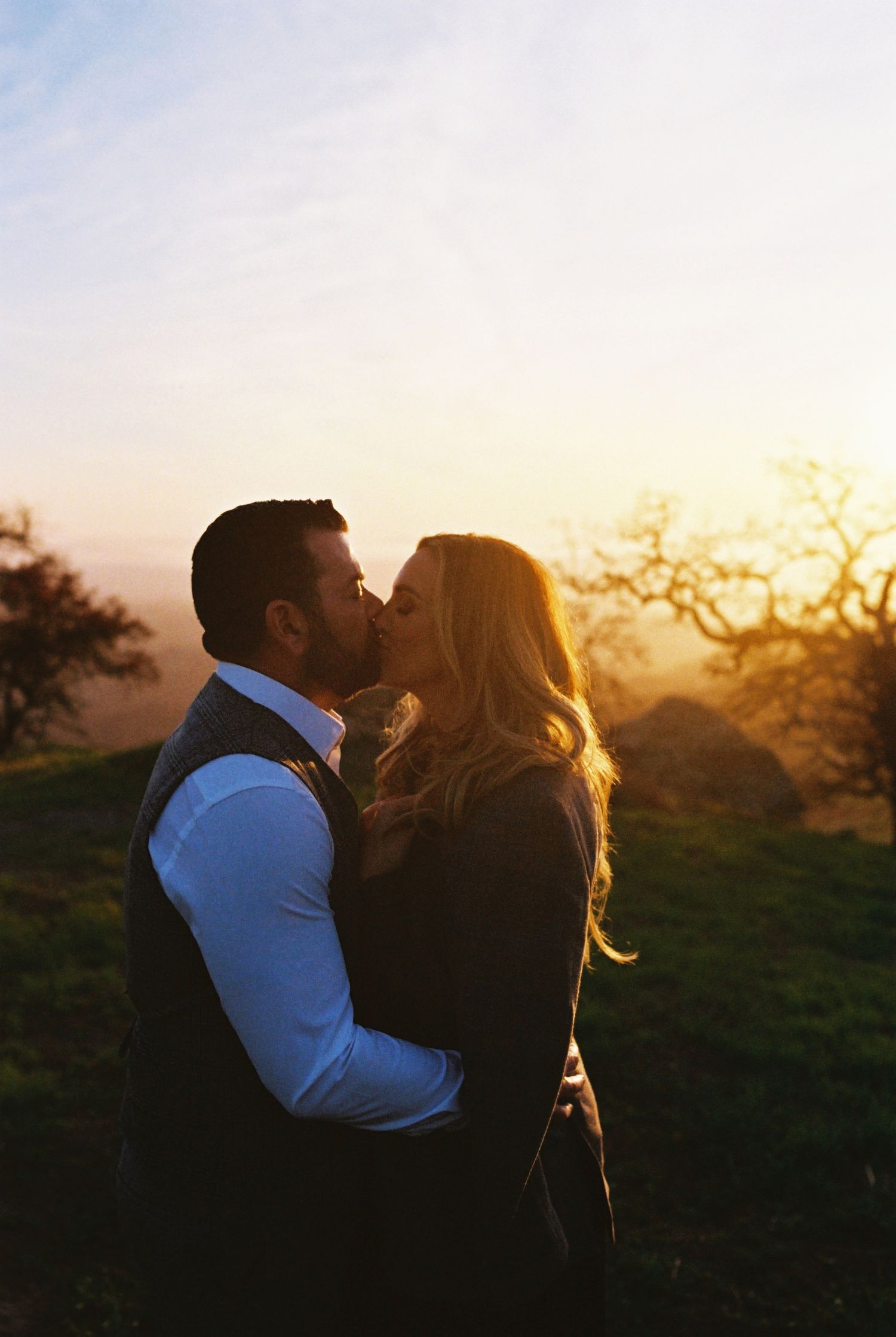 A couple shares a romantic kiss at sunset on a hilltop with golden light streaming through trees in the background.