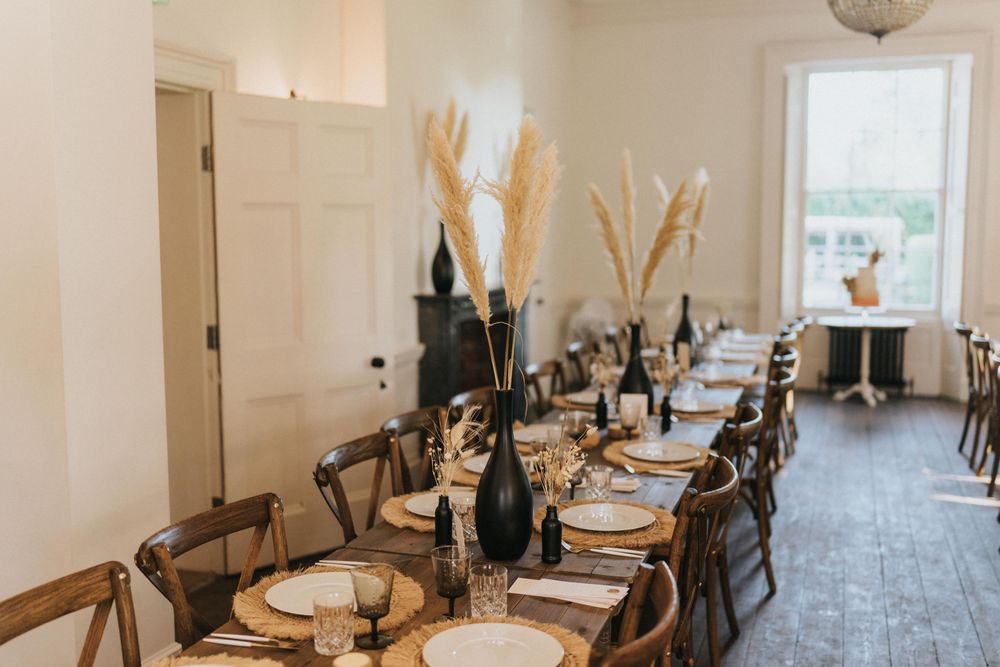A rustic dining table set for dinner features wooden chairs and pampas grass centrepieces in a bright room at Aswarby Rectory.