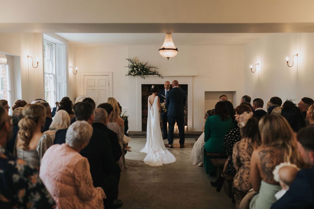 Indoor wedding ceremony takes place in an elegant white room with guests seated in rows watching the couple at the altar Aswarby Rectory.