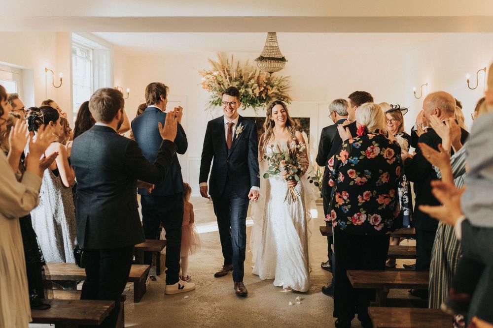 Elegant couple walks down the aisle while wedding guests take photos during ceremony in rustic indoor venue at Aswarby Rectory.