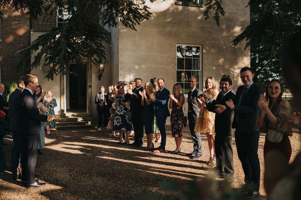 Wedding guests stand in a line clapping at an outdoor evening reception with golden sunlight streaming through trees at Aswarby Rectory.