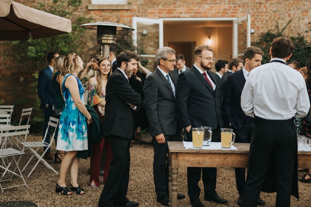 Wedding guests in formal attire gather in outdoor garden setting with string lights and brick walls creating ambiance at Aswarby Rectory.