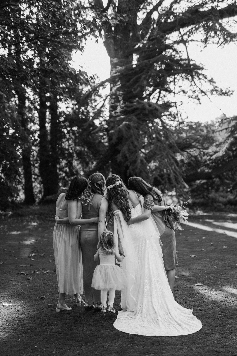 Black & white photo of a bridal party huddled together outdoors with bridesmaids in soft dresses and bride in white gown at Aswarby Rectory.
