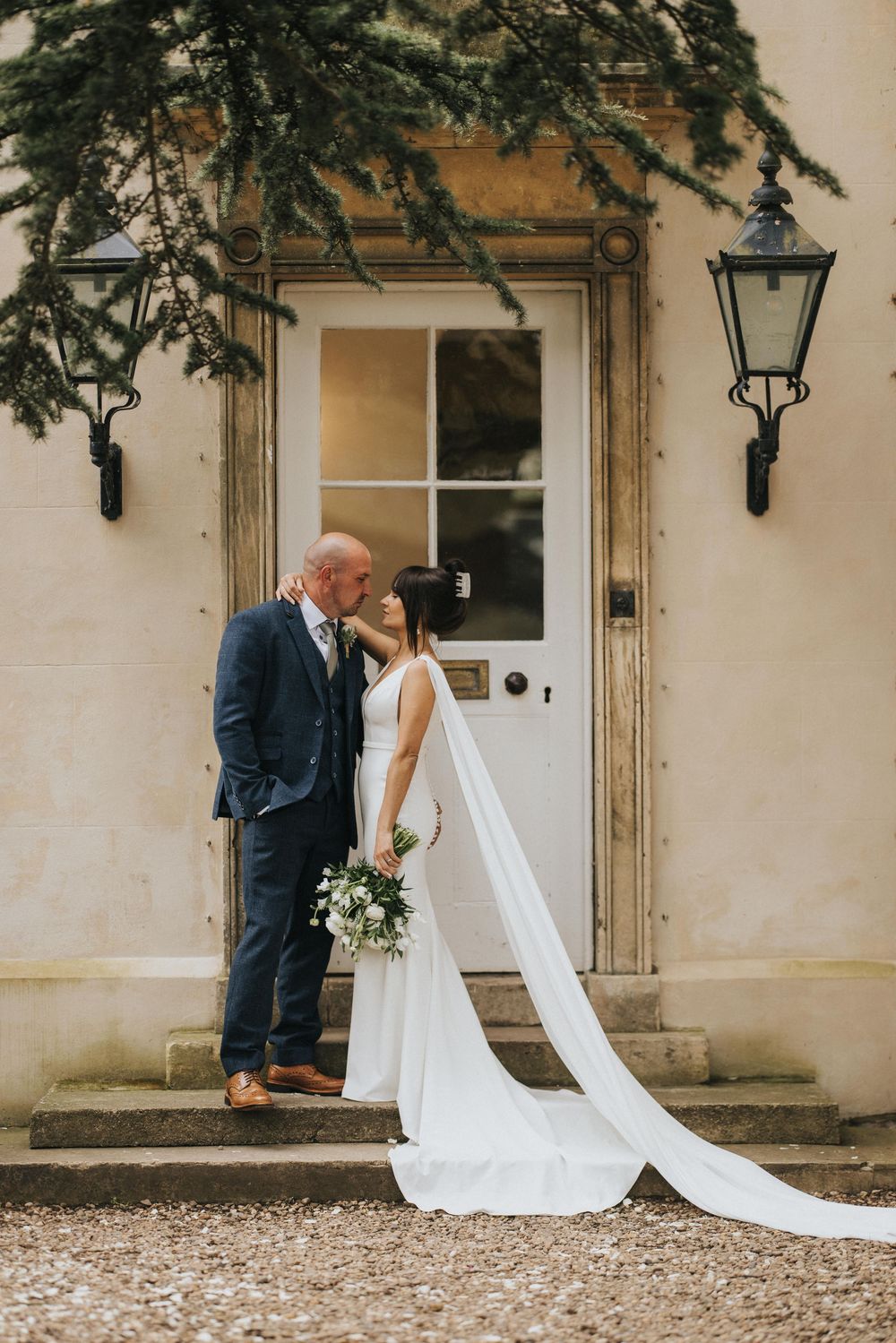 A couple in wedding attire share an intimate moment on stone steps in front of a white doorway with overhanging greenery at Aswarby Rectory.