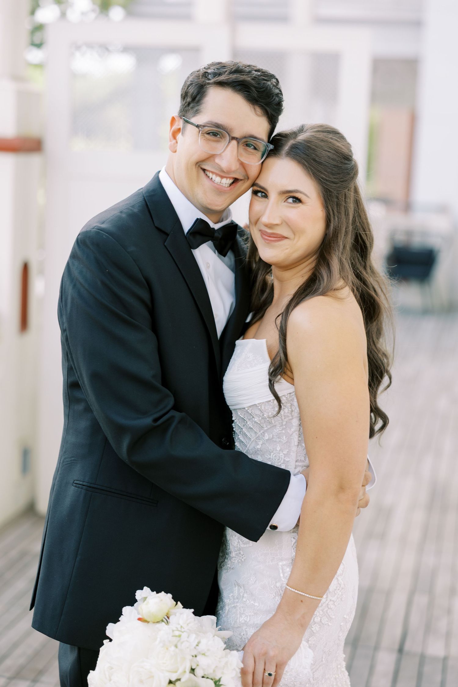 A romantic wedding couple poses together on a boardwalk in formal attire with white flowers and loving smiles.