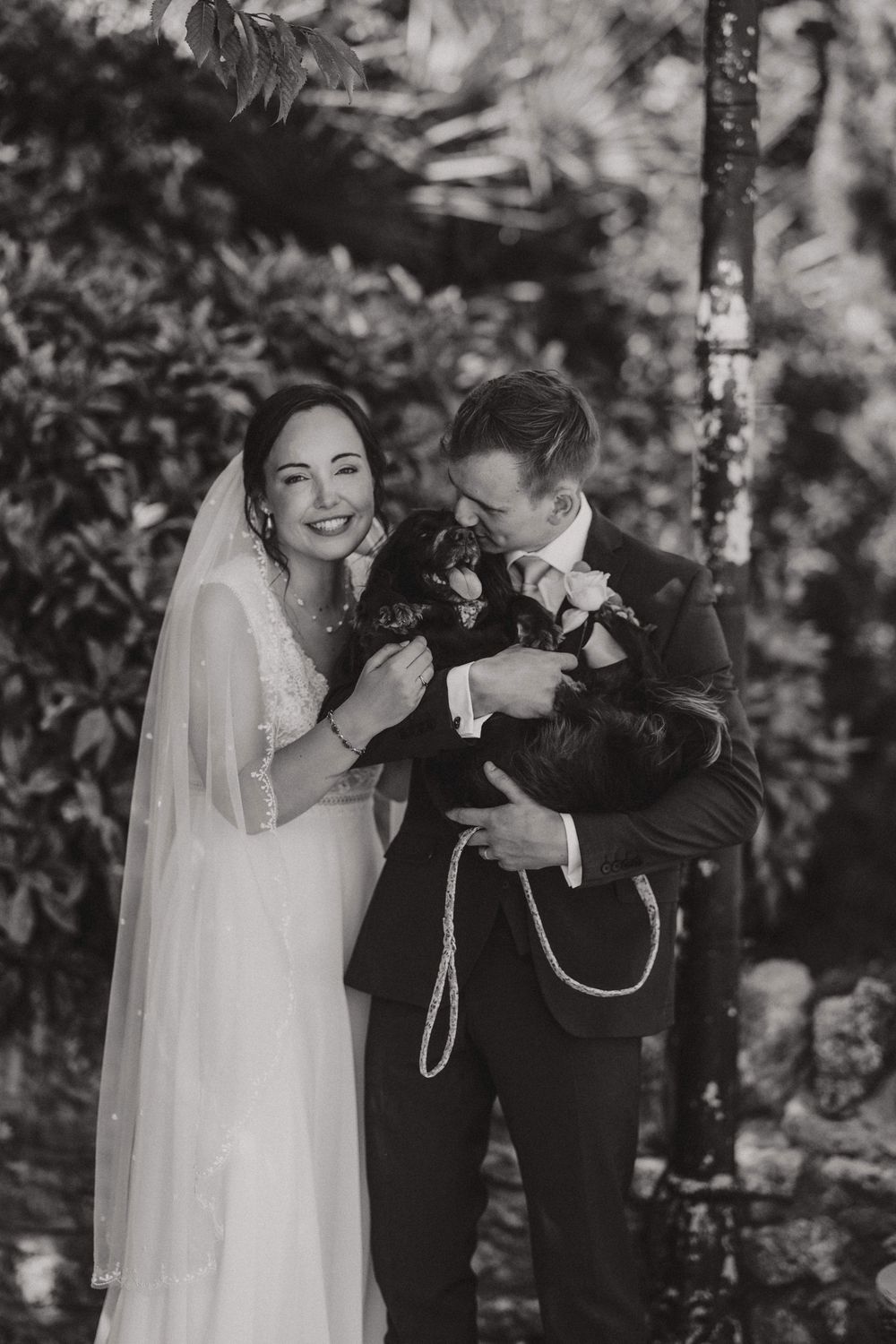A black and white wedding photo showing a bride and groom sharing a joyful moment together outdoors among trees.