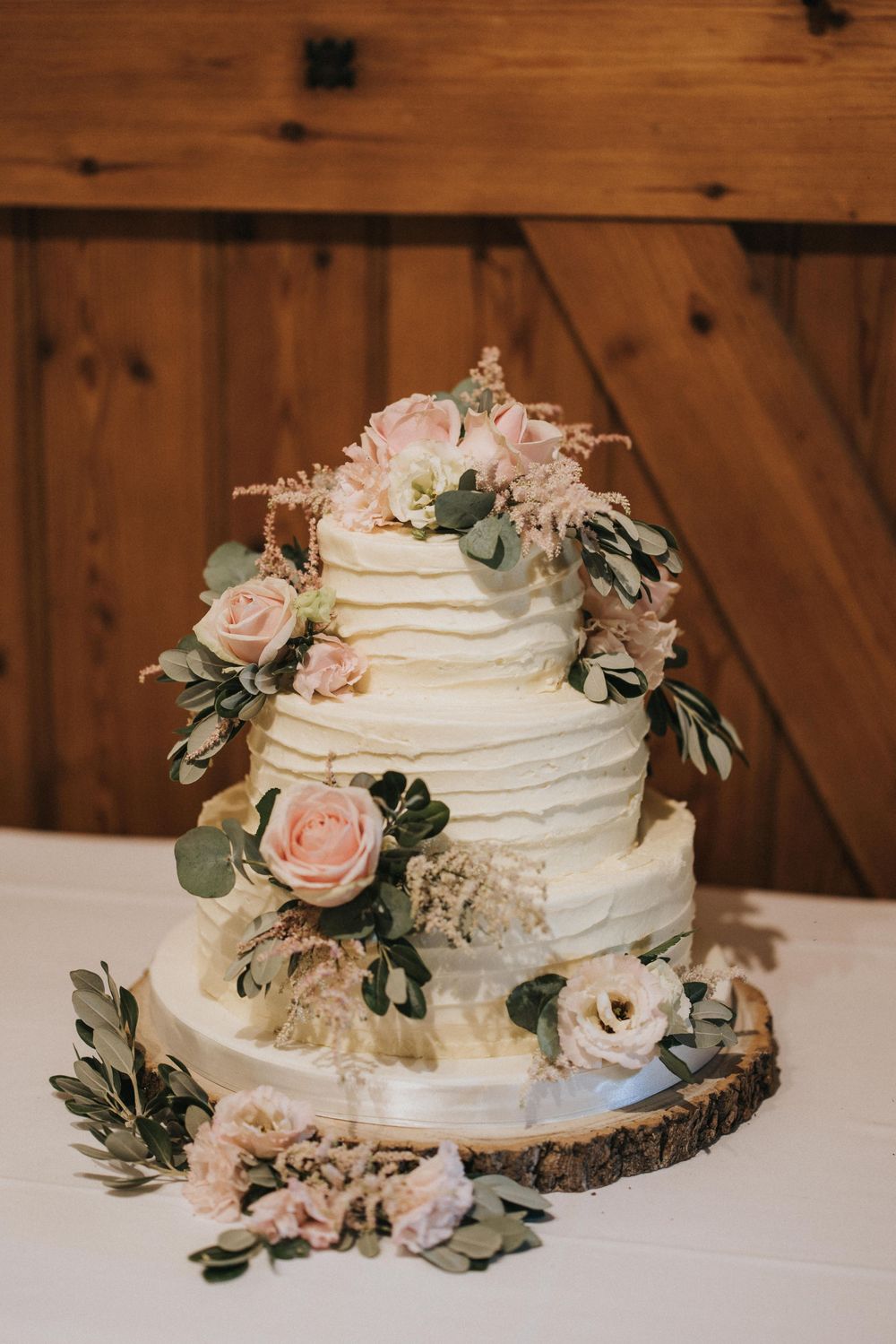 Elegant white wedding cake adorned with blush roses and eucalyptus on a rustic wood slice against a wooden backdrop.
