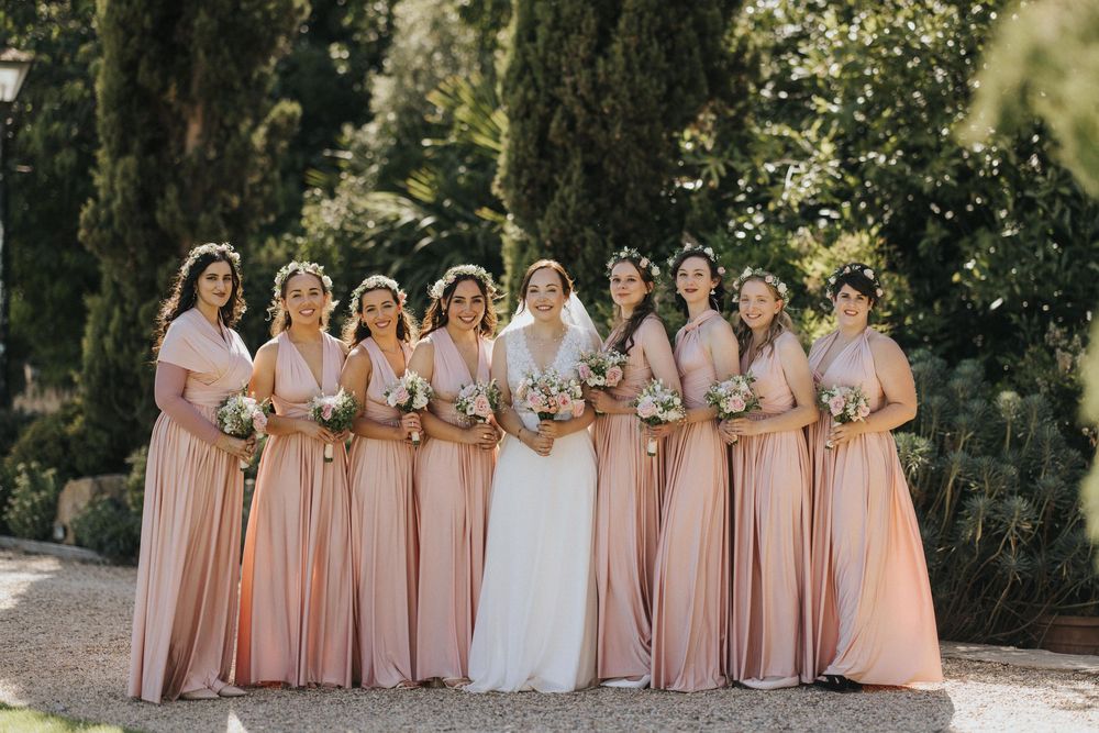 Wedding party wearing matching blush pink dresses stands outdoors among cypress trees on sunny day.