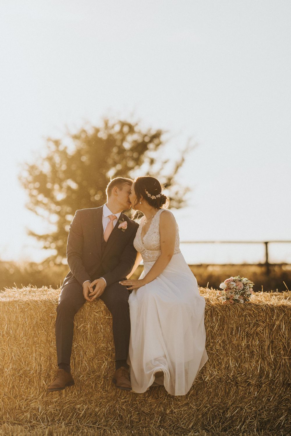 A couple sits together on hay bales at sunset during a rustic outdoor wedding ceremony.