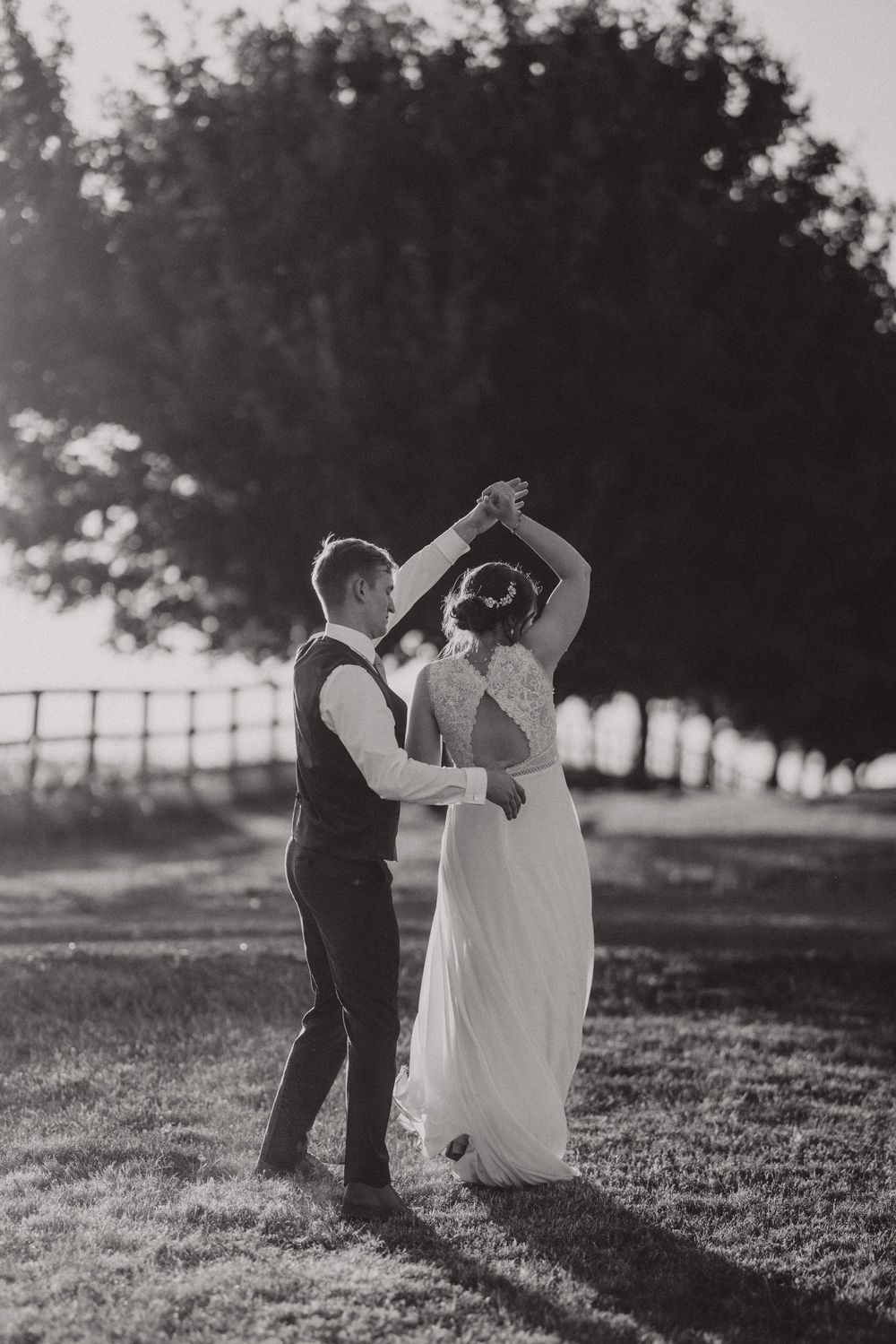 A couple in wedding attire dances together on a grassy hill during sunset, creating a romantic black and white silhouette.