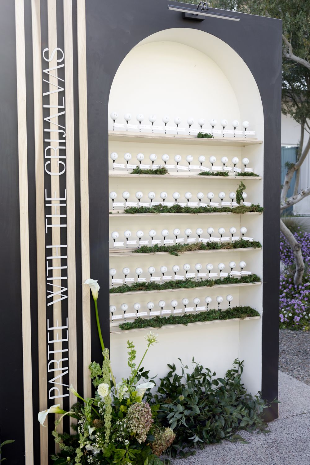 Modern black and white escort card display with multiple shelves in an arched design and floral accents.
