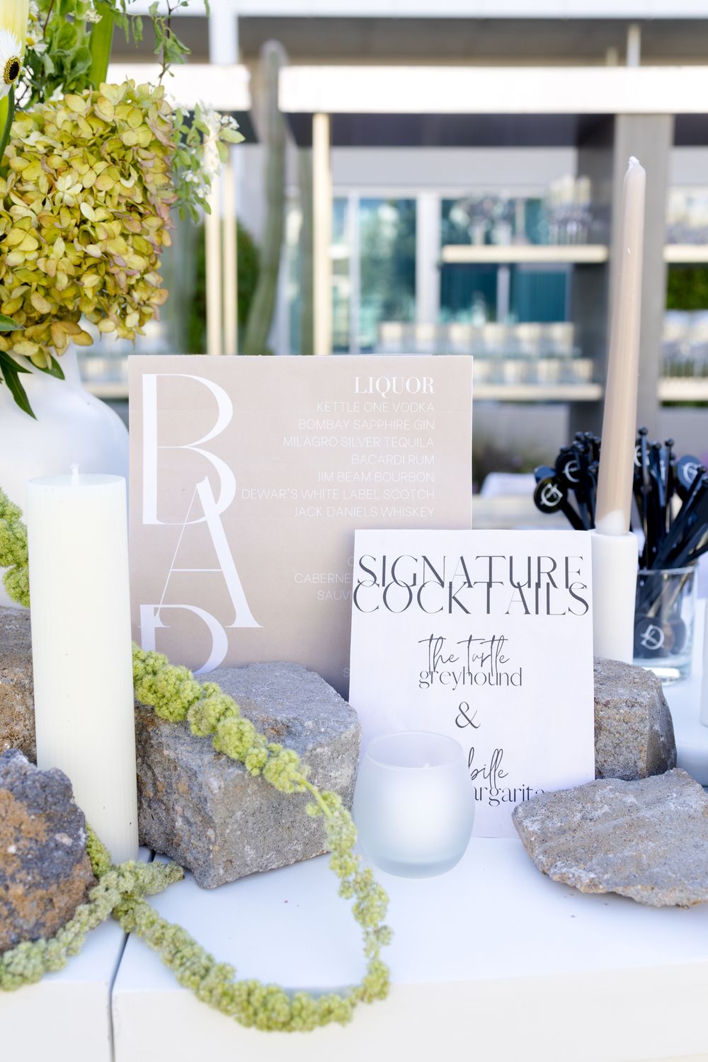 Elegant wedding signage for signature cocktails displayed with white candles, green hydrangeas and decorative stones.