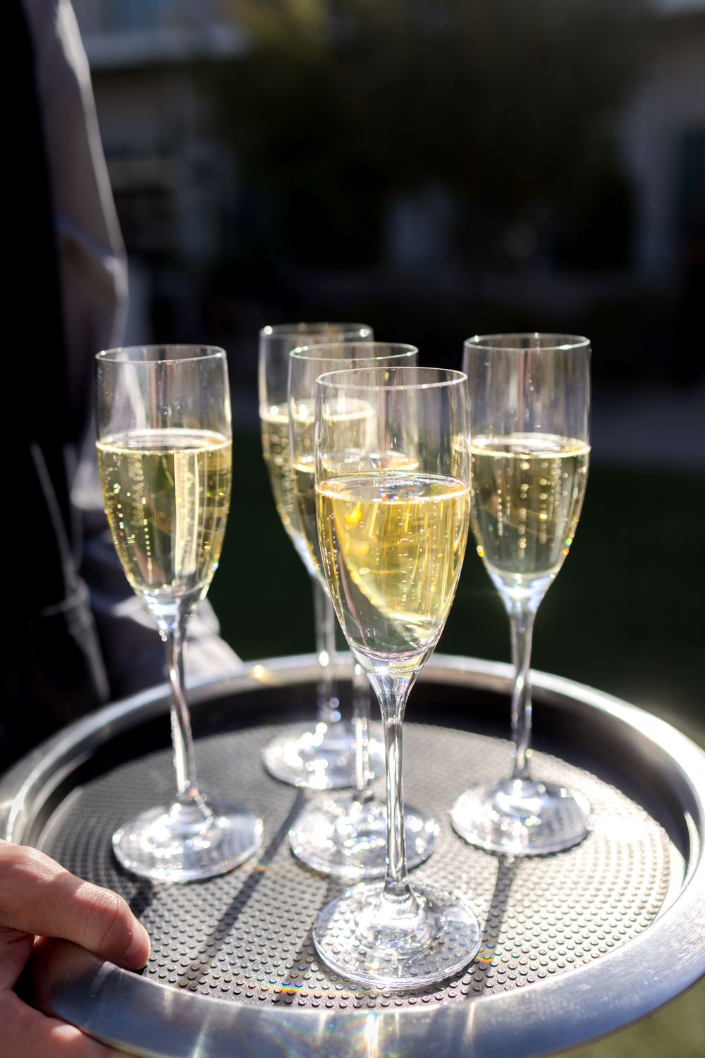 A silver serving tray holds multiple elegant stemmed wine glasses filled with golden champagne in sunlight.