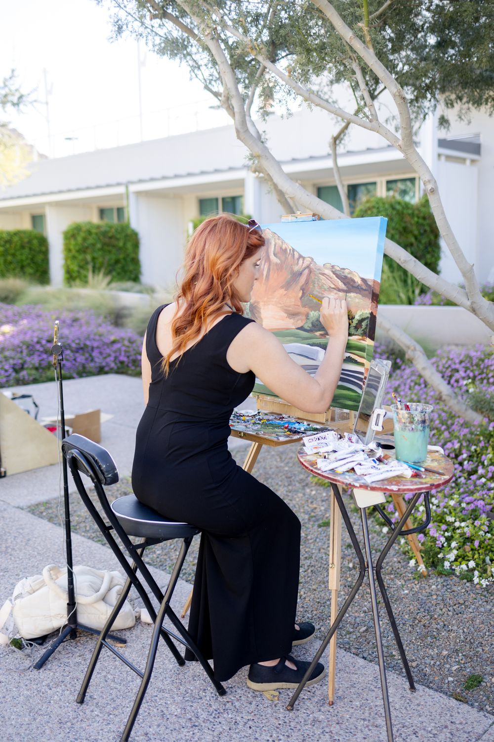 Artist working on watercolor painting outdoors with art supplies on small table in garden setting.