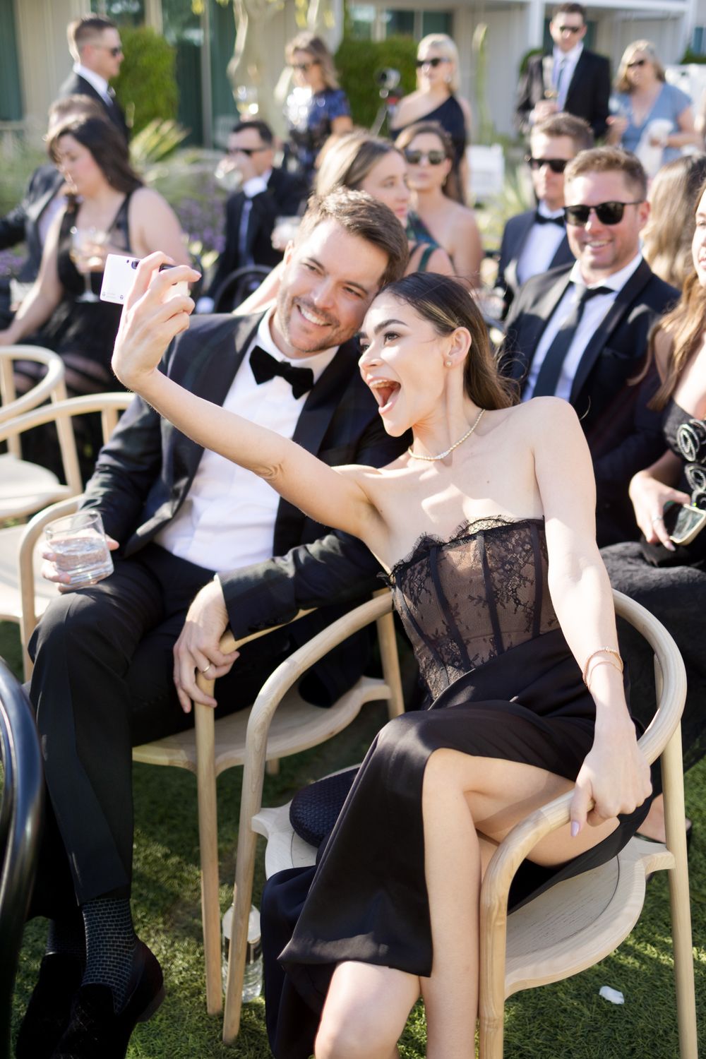 A couple in formal black attire laughs while taking a selfie at an outdoor event with other guests seated nearby.