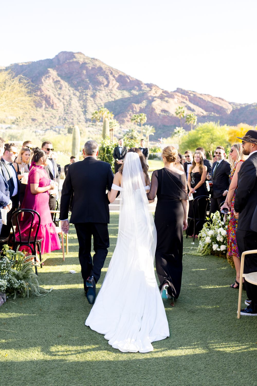 Outdoor wedding guests gather on a grass lawn with desert mountains in the backdrop during a sunny daytime ceremony.