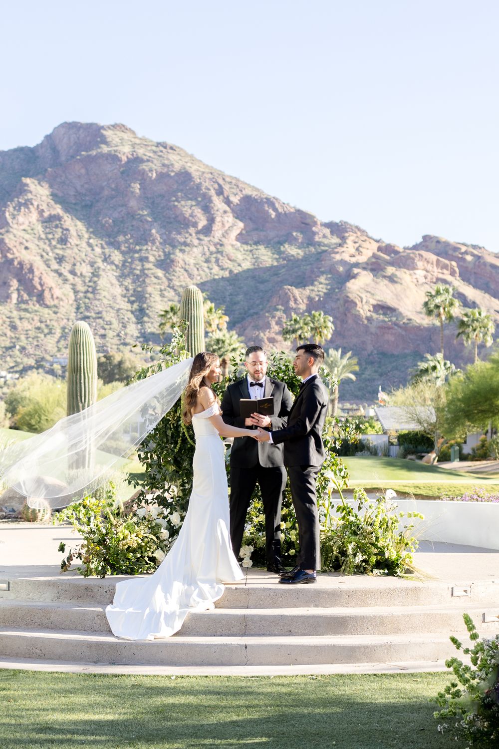 Bride's white veil flows in the wind against a majestic desert mountain backdrop during an outdoor wedding ceremony.