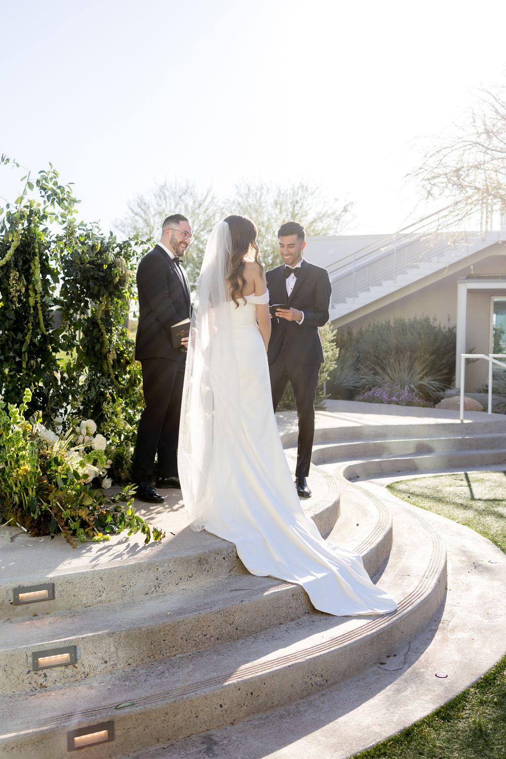 Wedding ceremony taking place on curved outdoor steps with flowing white dress in sunlit garden setting.