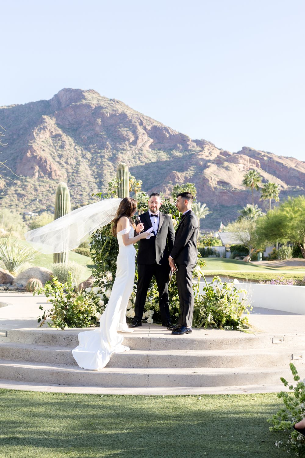 An outdoor wedding ceremony takes place on stone steps with desert mountains and cacti in the background.