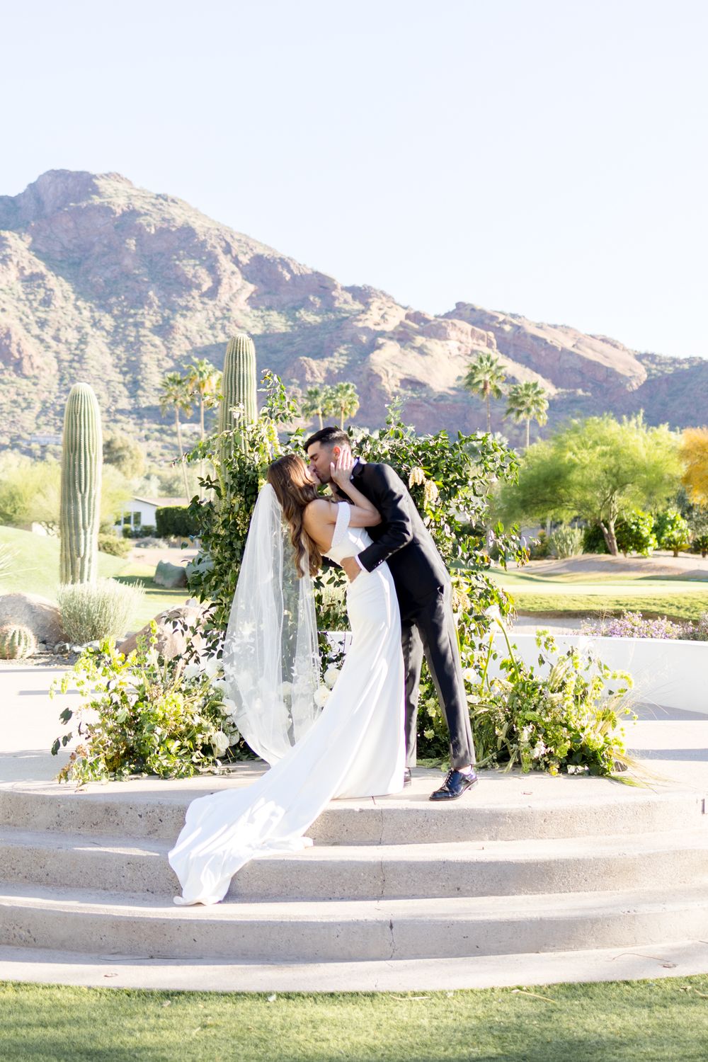 A wedding couple shares a romantic kiss on stone steps with desert mountains and saguaro cacti in the background.