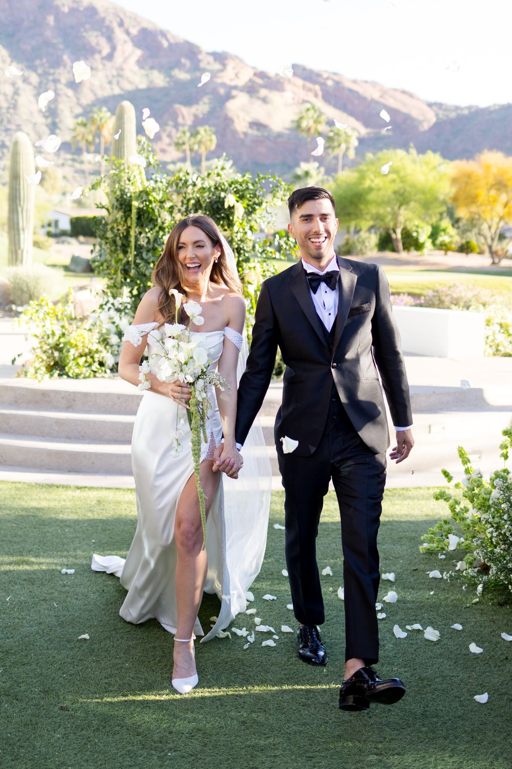 A wedding couple walks hand in hand along a garden path with desert mountains and cactus in the background.