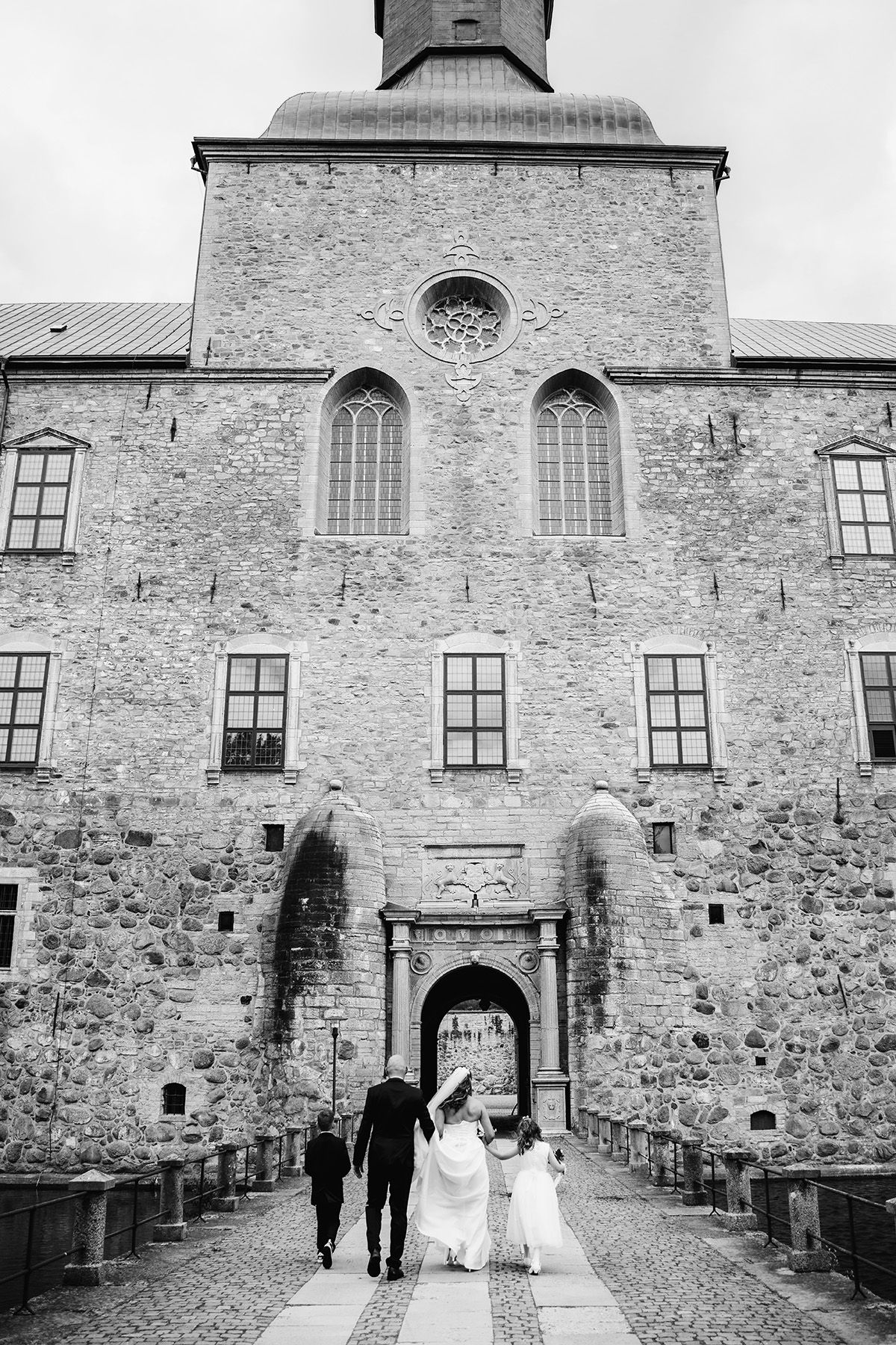 Bröllop i Vadstena, bröllopsporträtt utanför Vadstena slott. Brudparet går med sina barn över bron, foto av bröllopsfotograf i Östergötland.