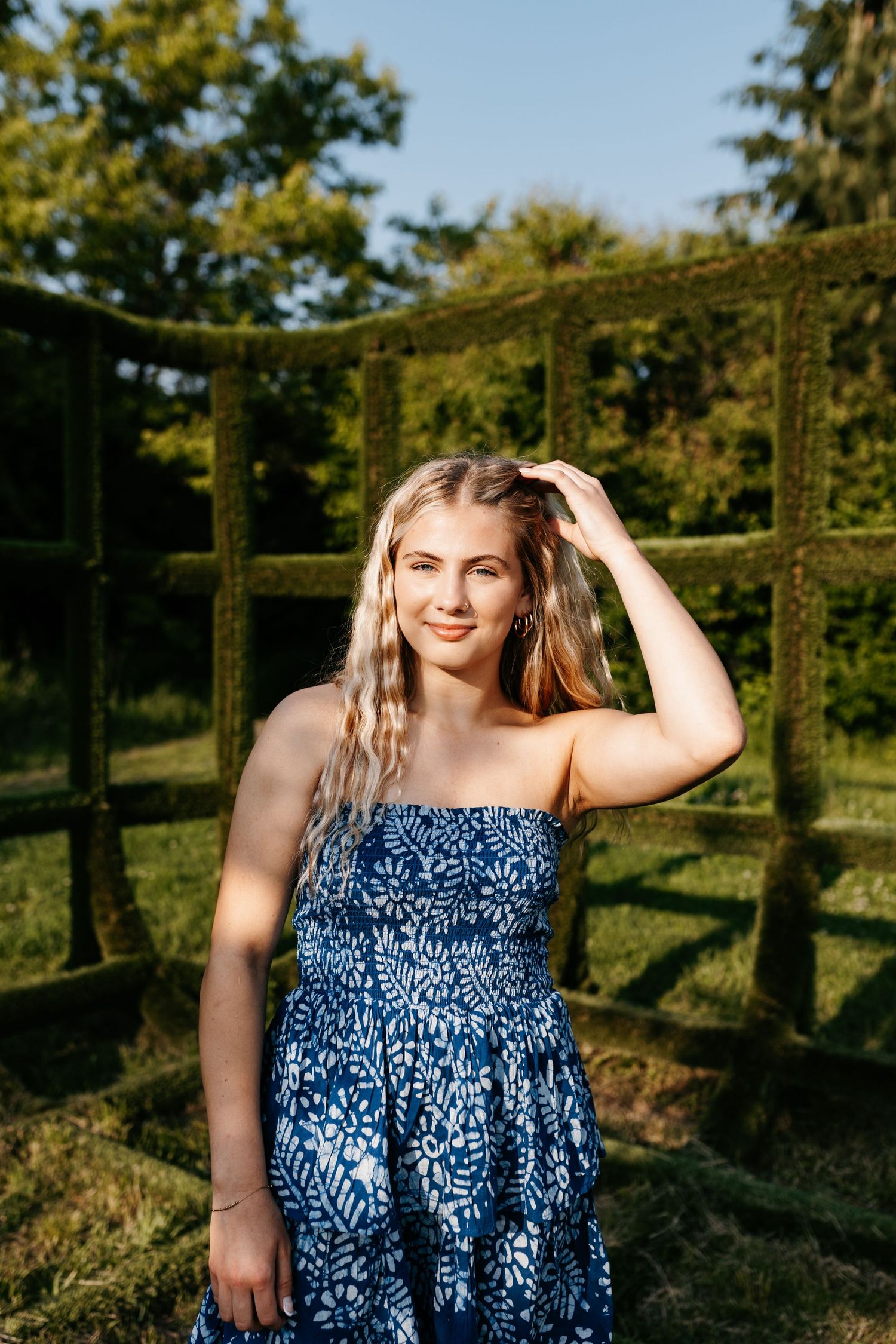 Woman wearing blue and white floral print dress poses in sunlit garden setting with trees and greenery in background.