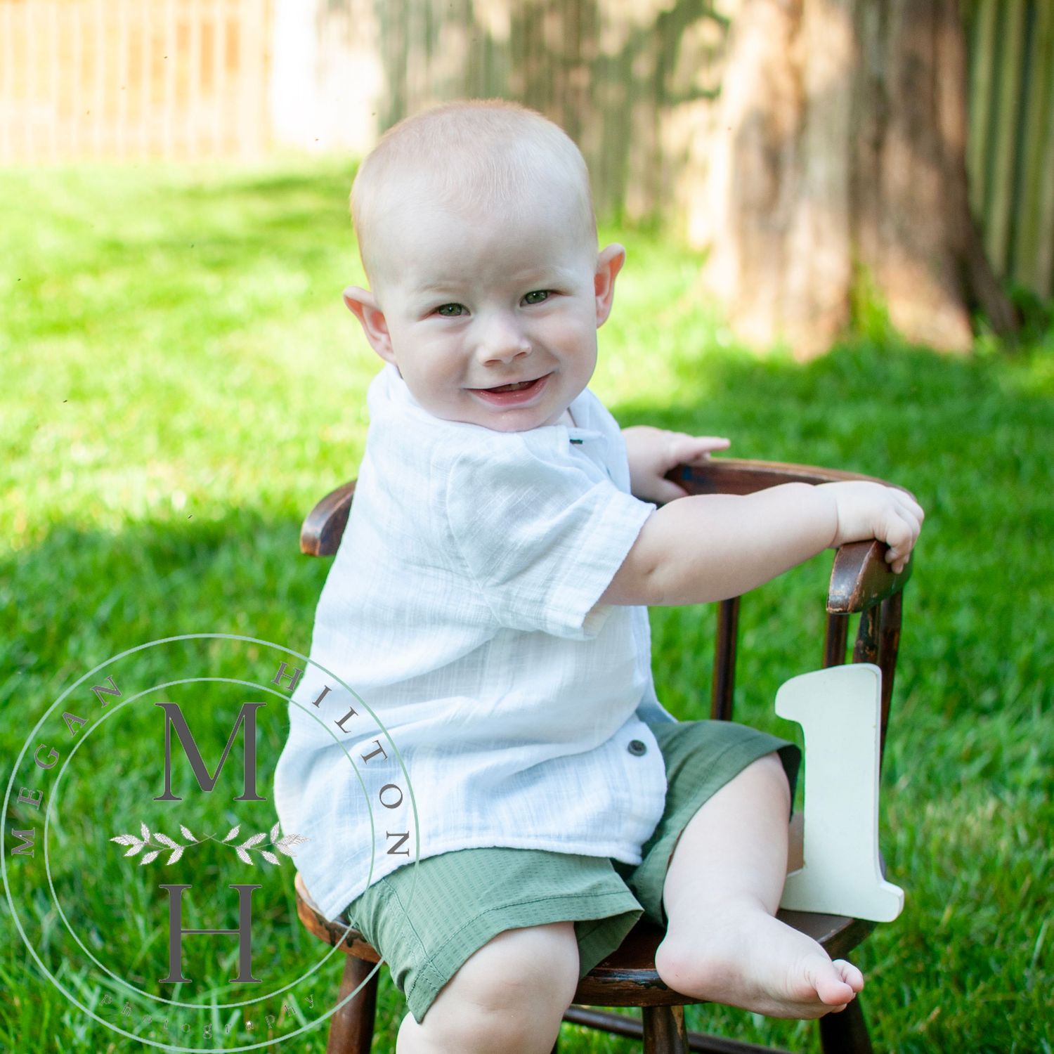 Small child in white shirt and green shorts sitting on a wooden chair outdoors on grass in sunlight.