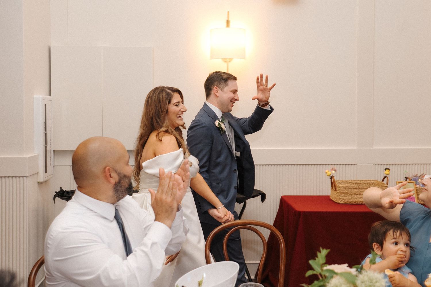 People celebrating at a restaurant event with tables and chairs, as participants wave and interact.