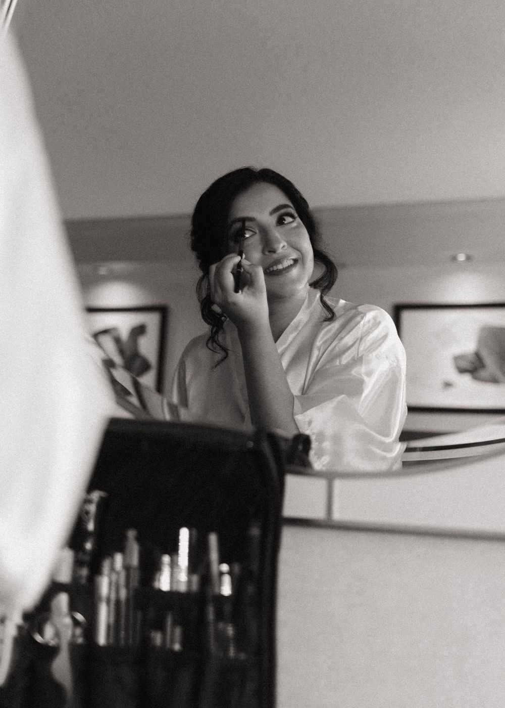 Black and white photo showing someone in a white silk robe applying makeup in front of a mirror at a vanity table.