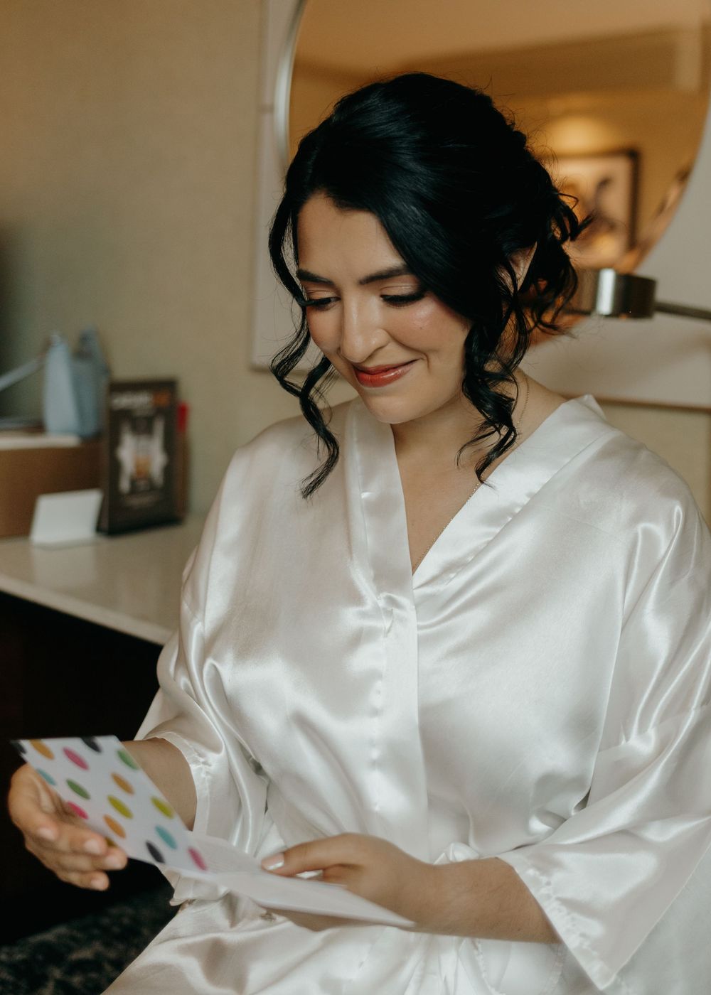 Woman in white silk robe smiles while looking down at colorful greeting card in natural light setting.