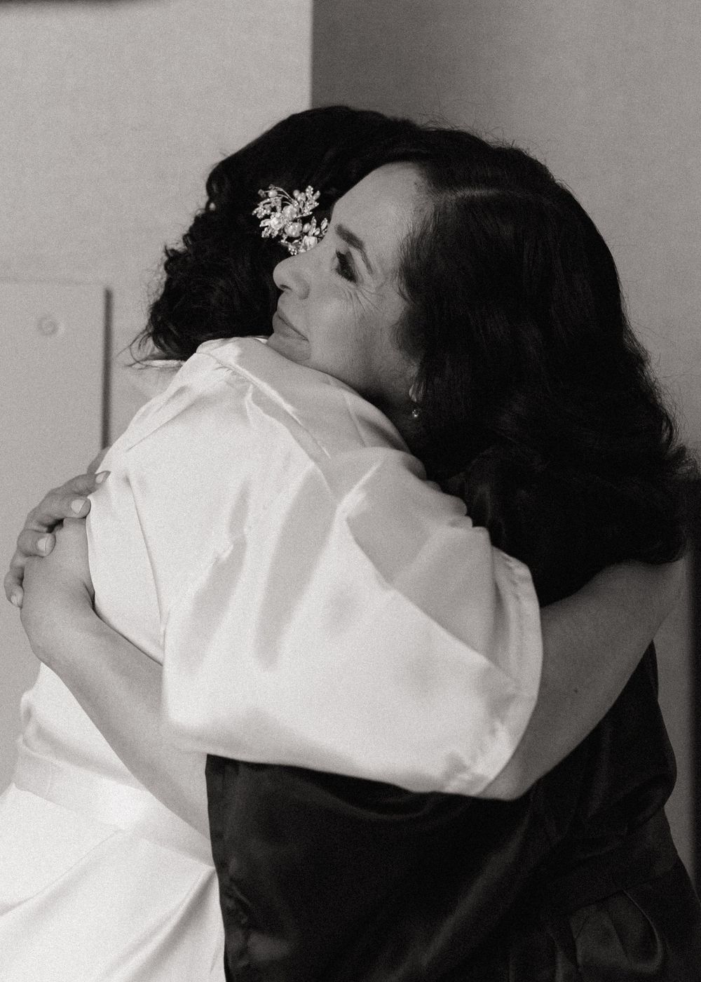 Black and white photograph of a tender embrace between two people in formal attire with delicate hair accessories.