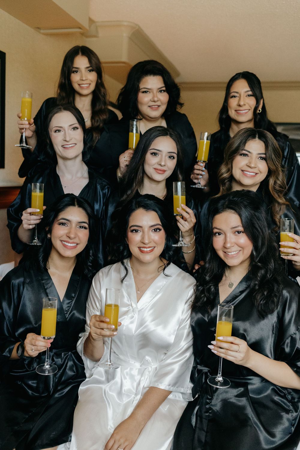 Bridesmaids in black silk robes and bride in white robe hold up mimosa glasses for a celebratory pre-wedding photo.