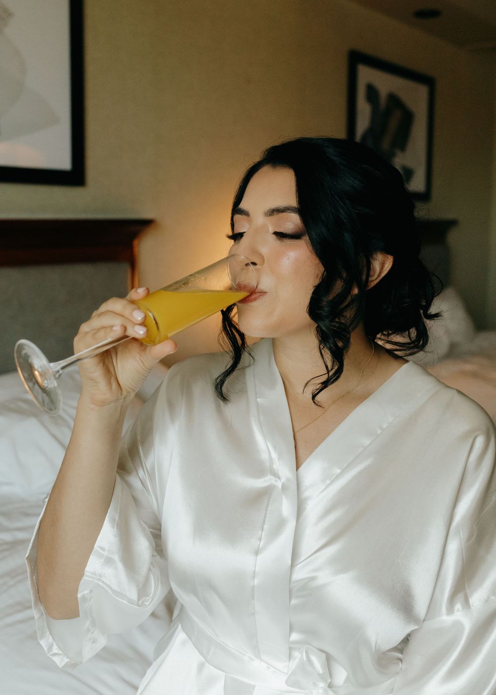 A person in a white silk robe sips from a glass of champagne or mimosa in what appears to be a bedroom setting.
