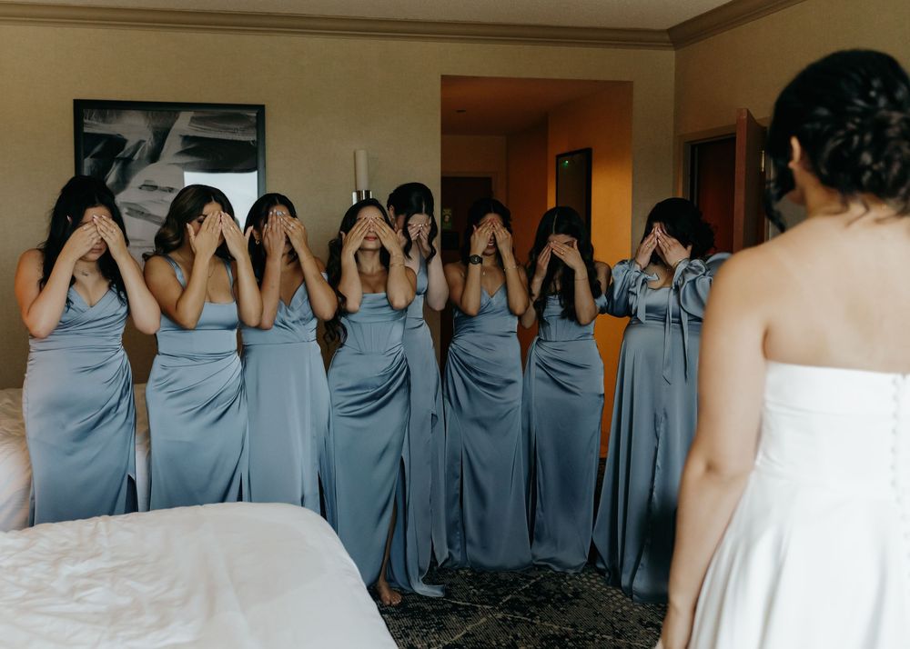 Bridesmaids in light blue dresses cover their eyes while looking at someone wearing a white dress in a hotel room.