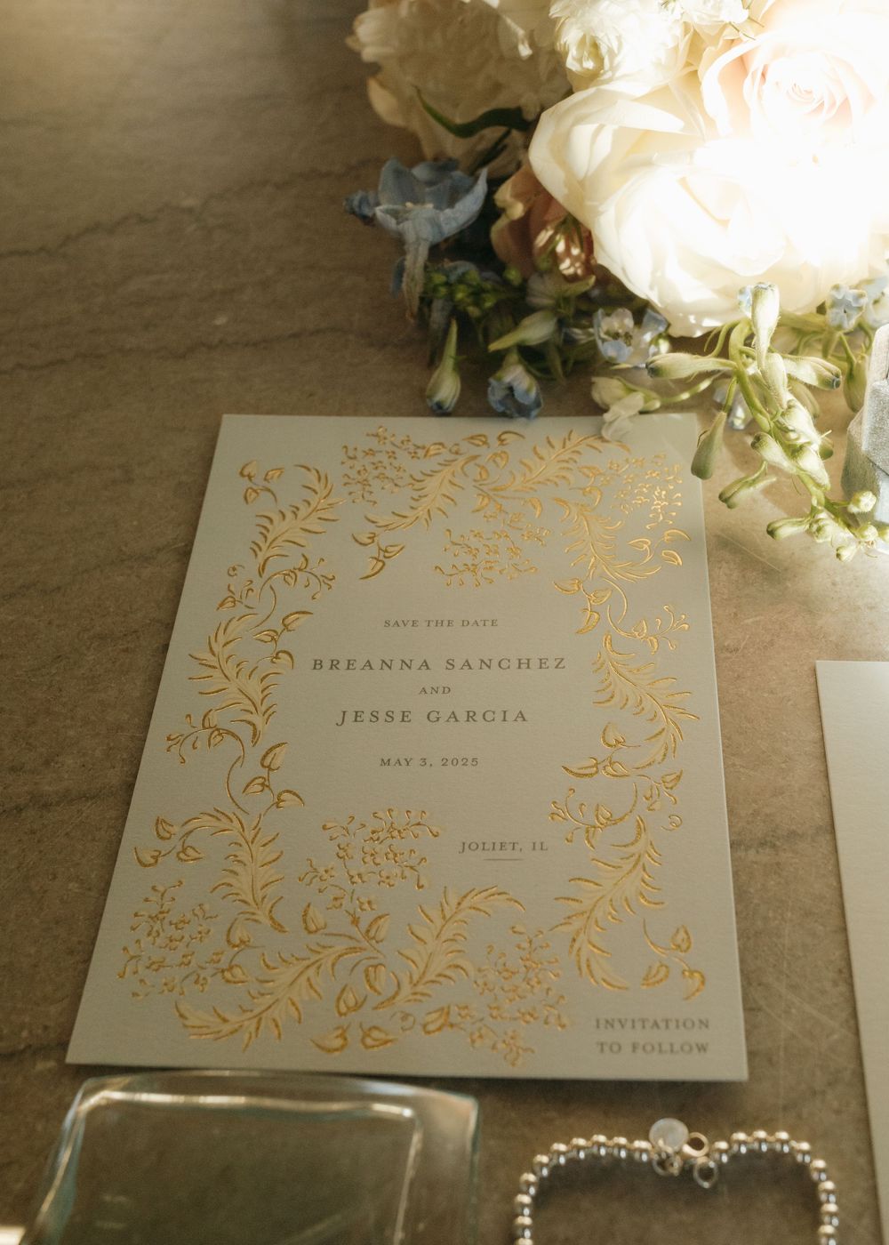 Gold-foiled wedding invitation with floral border design surrounded by white roses and pearl jewelry.