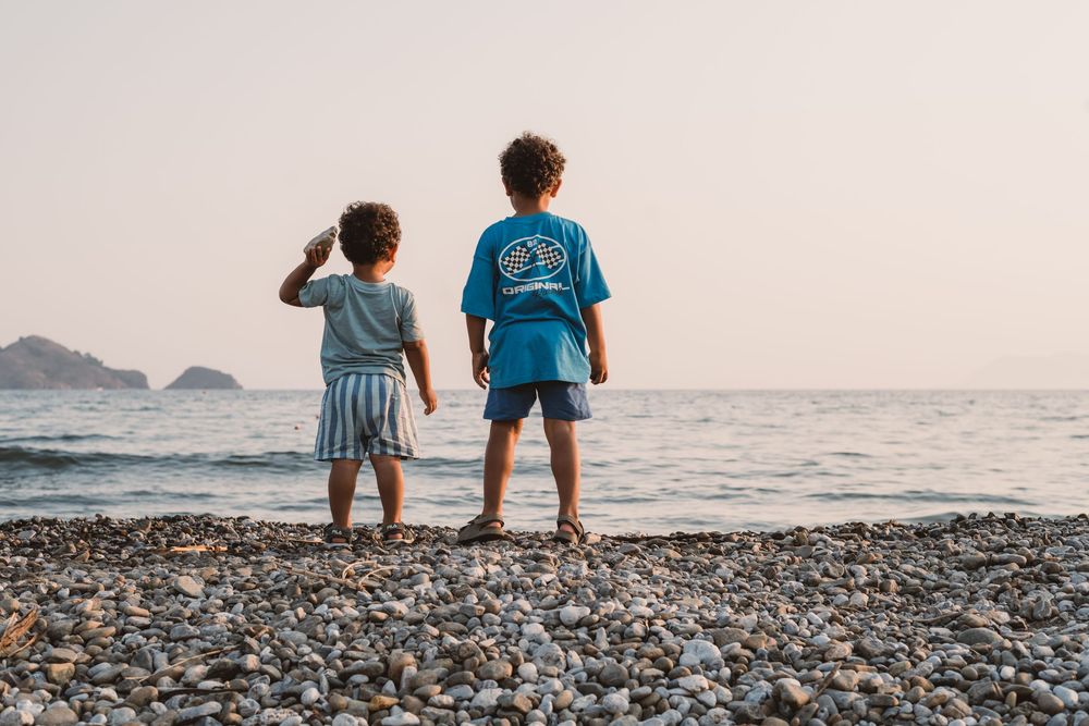 Silhouettes of children sitting on a rocky beach at sunset with mountains visible in the distance.