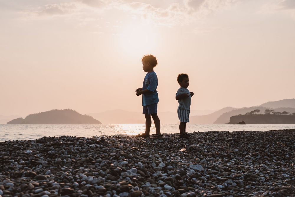 Two silhouetted children stand on rocky beach at sunset with mountains in background and moody pink sky.